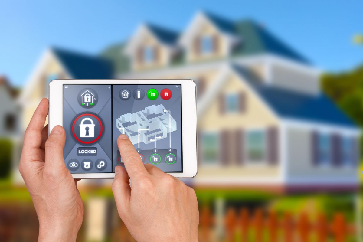 Hands holding a tablet displaying a home security system, with a home security setup visible in the background.