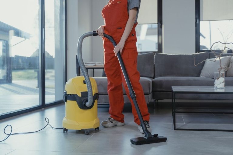 A woman in an orange overall stands beside a vacuum cleaner, ready to clean the room.